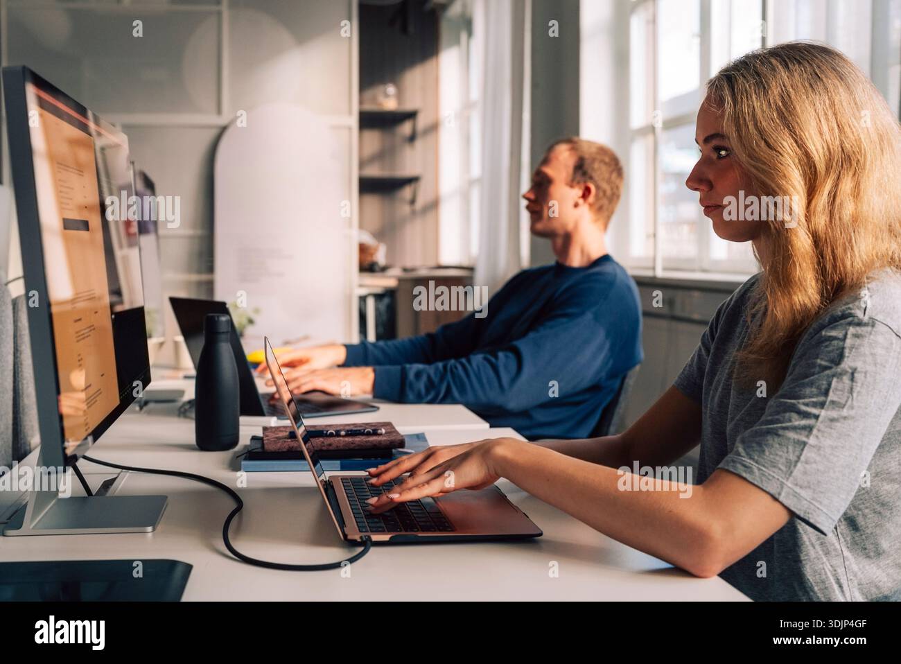 Blond female programmer coding on laptop and computer while sitting at desk in startup office Stock Photo