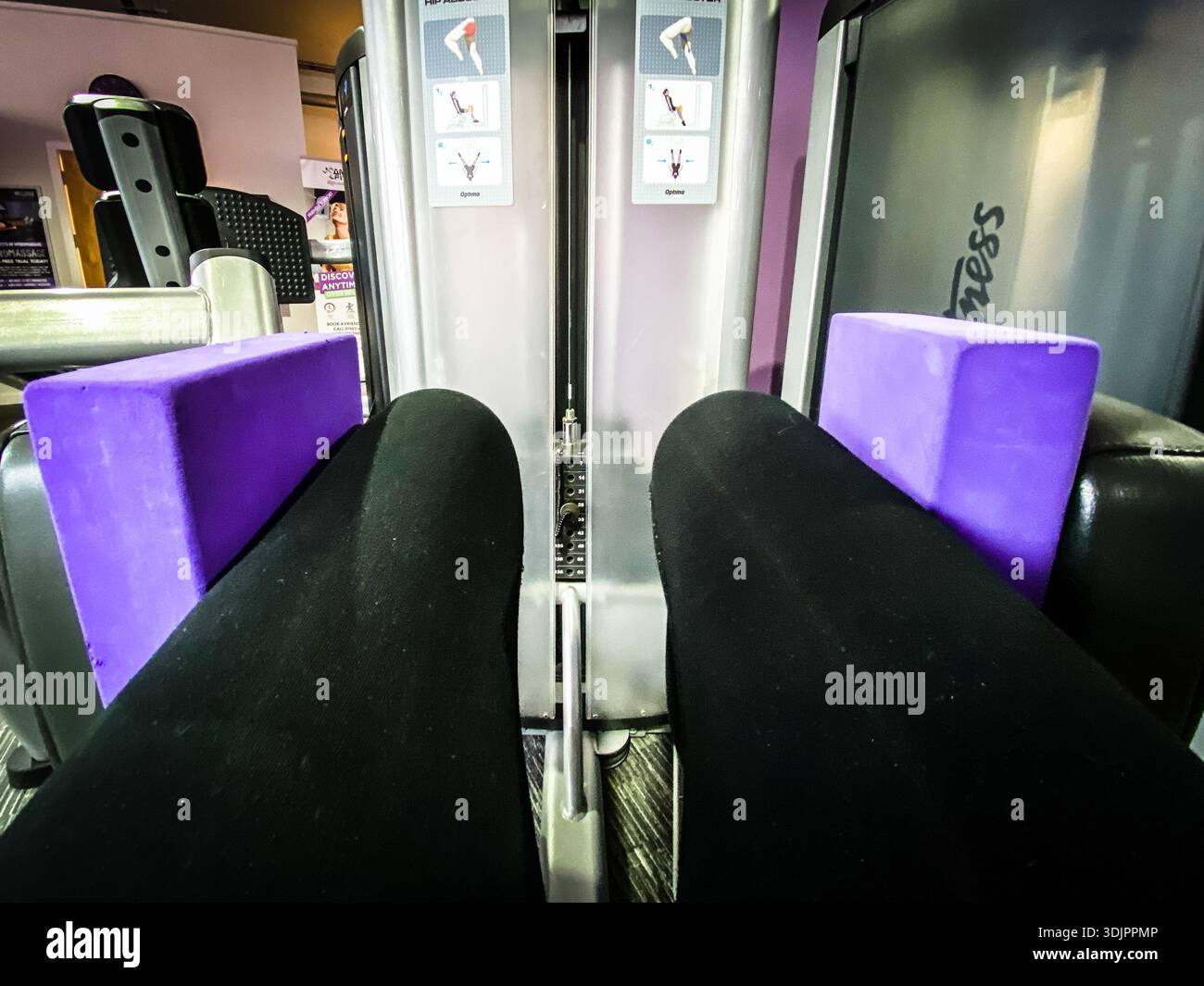 Closeup view from the seat of a hip abductor gym machine showing a woman’s legs in black leggings between purple pads at an indoor fitness club in the - Smartphone Captured Stock Image