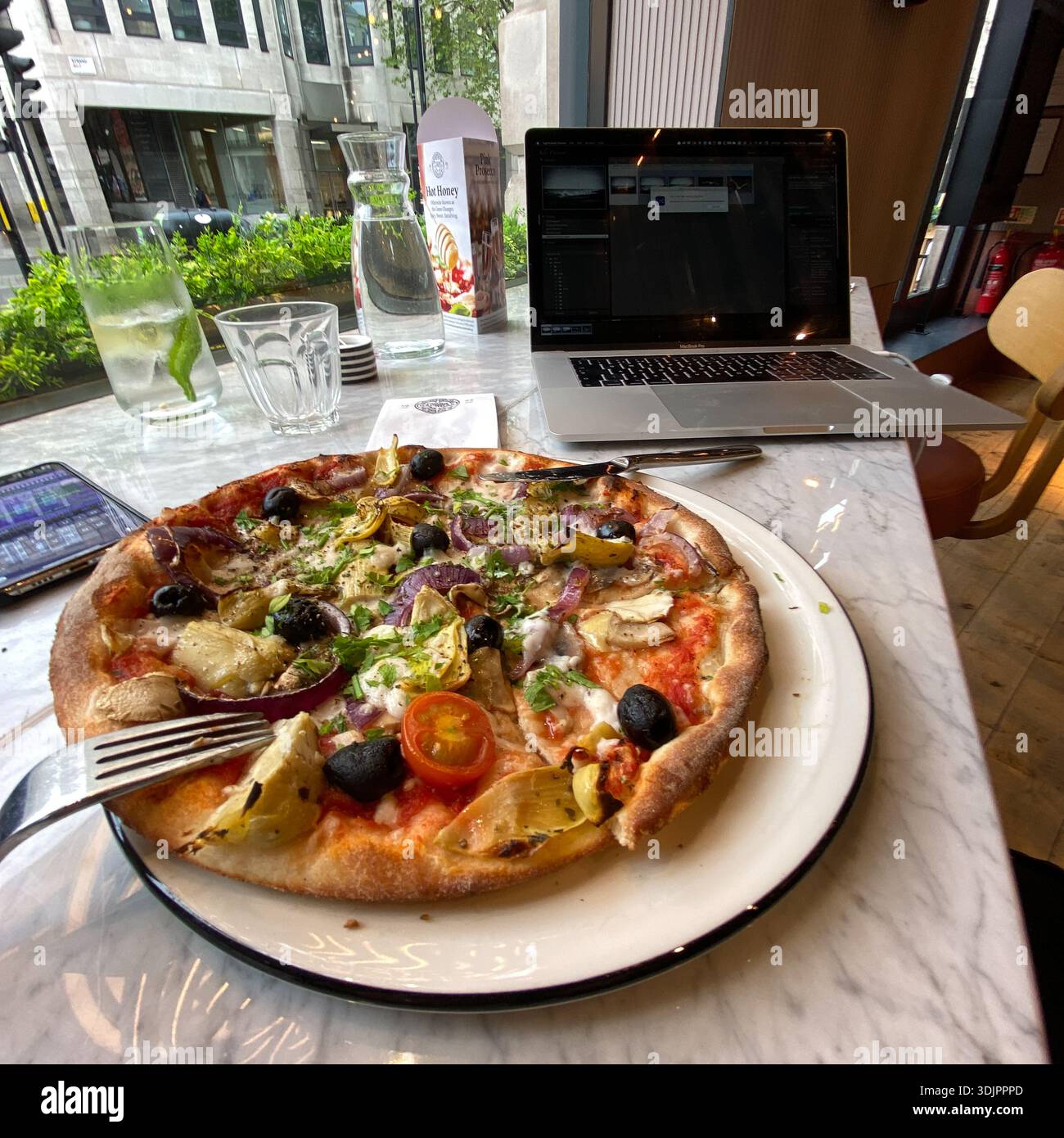 Pizza and drinks on a marble table beside a laptop in a London restaurant, illustrating a casual remote working lunch with a city street view through - Smartphone Captured Stock Image