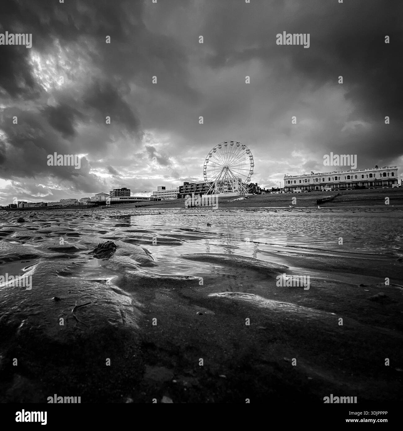Stormy skies over Worthing seafront in West Sussex, with the observation wheel and promenade buildings reflected in wet sand at low tide in a moody bl - Smartphone Captured Stock Image