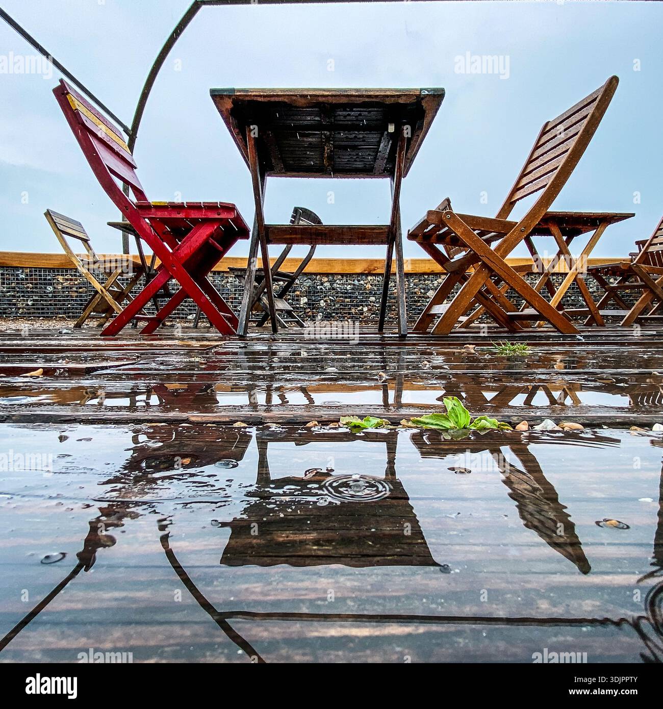 Wet wooden table and chairs on a deserted seaside terrace after rain, with puddles reflecting the structure against a cloudy sky in England. - Smartphone Captured Stock Image