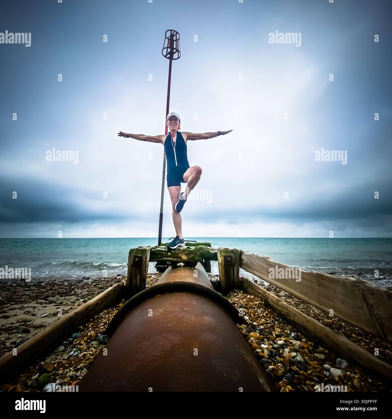 Woman practising tree pose yoga on a rusty outfall pipe amid pebbles on an English beach, against a cloudy sea horizon - Smartphone Captured Stock Image