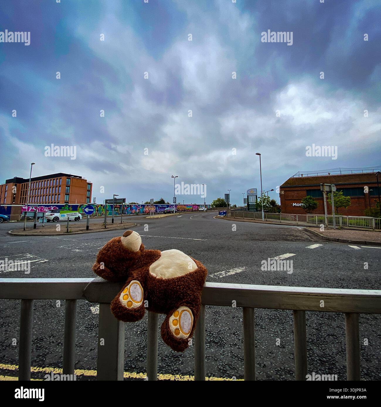 Lonely teddy bear draped over a barrier rail on an empty urban roundabout near modern buildings and graffiti murals under a cloudy sky in England - Smartphone Captured Stock Image