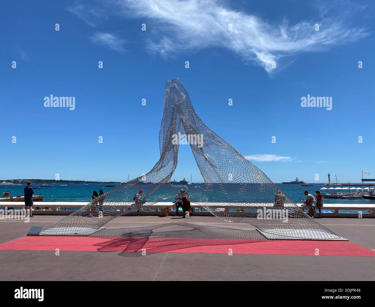 Large metallic mesh sculpture resembling a flowing dress on the promenade in Cannes, France, with sea, yachts and passers-by under a blue summer sky - Smartphone Captured Stock Image