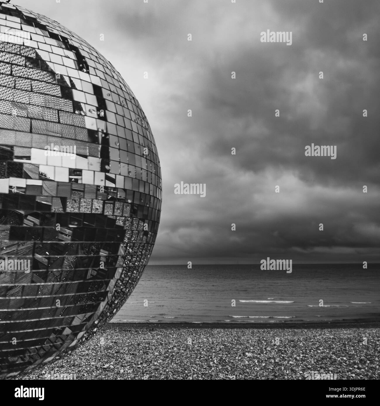 Large mirrored disco ball sculpture on a shingle beach with stormy skies and sea horizon, rendered in dramatic black and white - Smartphone Captured Stock Image