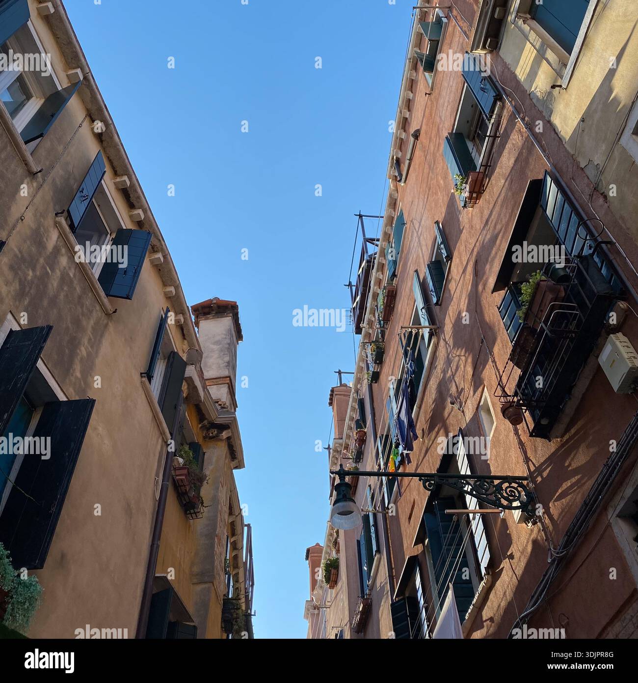 Upward view of narrow Venetian alleyway flanked by terracotta ochre buildings with green shutters, flower boxes, laundry lines and ornate street lamp - Smartphone Captured Stock Image