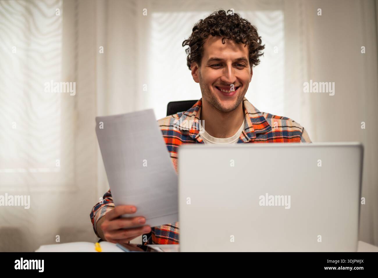 Cheerful young freelancer using laptop and checking documents while working at his cozy home. Stock Photo
