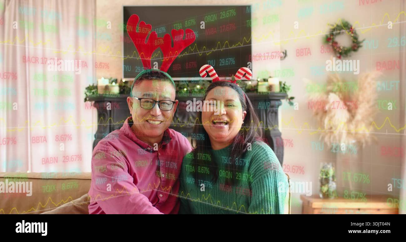 Laughing couple wearing novelty headbands and sweaters on sofa at home with stock market overlay Stock Photo