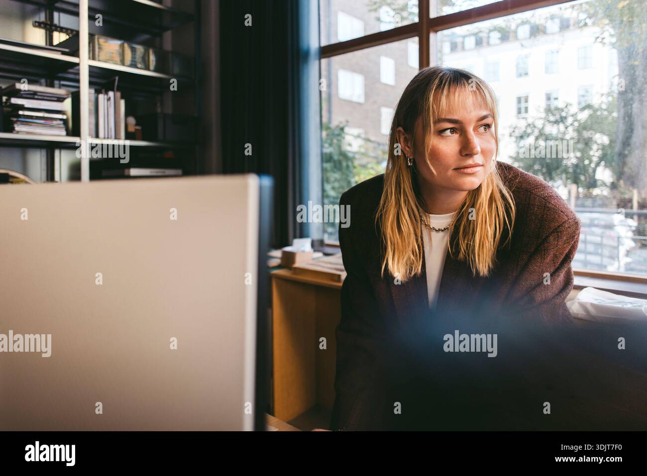 Young female creative professional looking away while leaning on desk in office Stock Photo