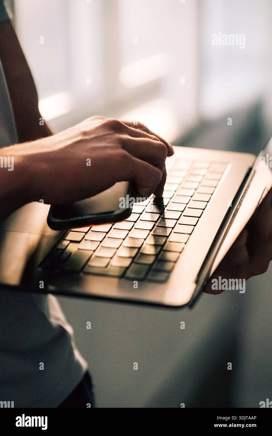 Hand of businessman holding smart phone while pressing key on laptop Stock Photo