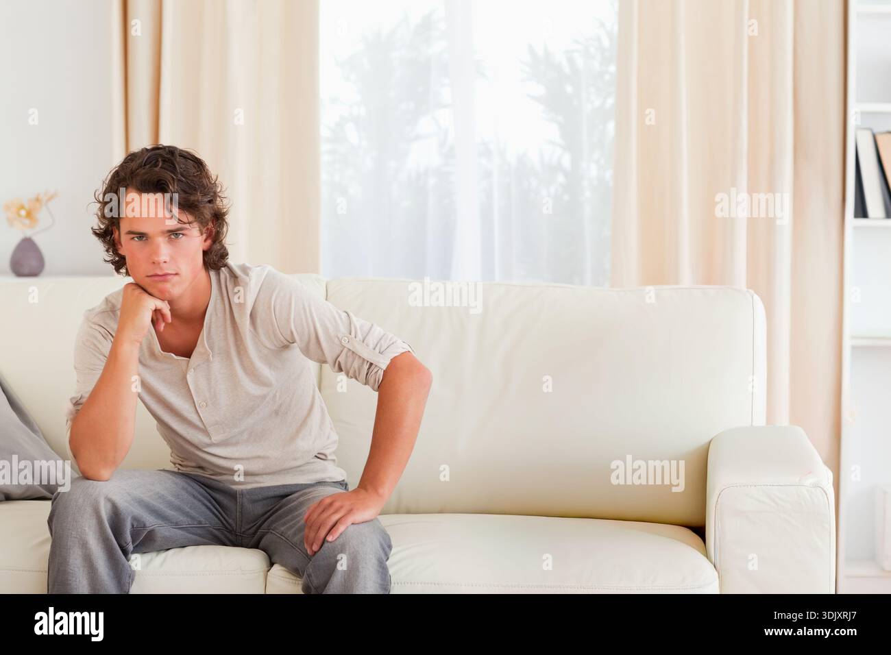 Man sitting on white leather sofa in living room, wearing beige henley and looking toward camera. Person, couch, interior, minimal, neutral, daylight, Stock Photo