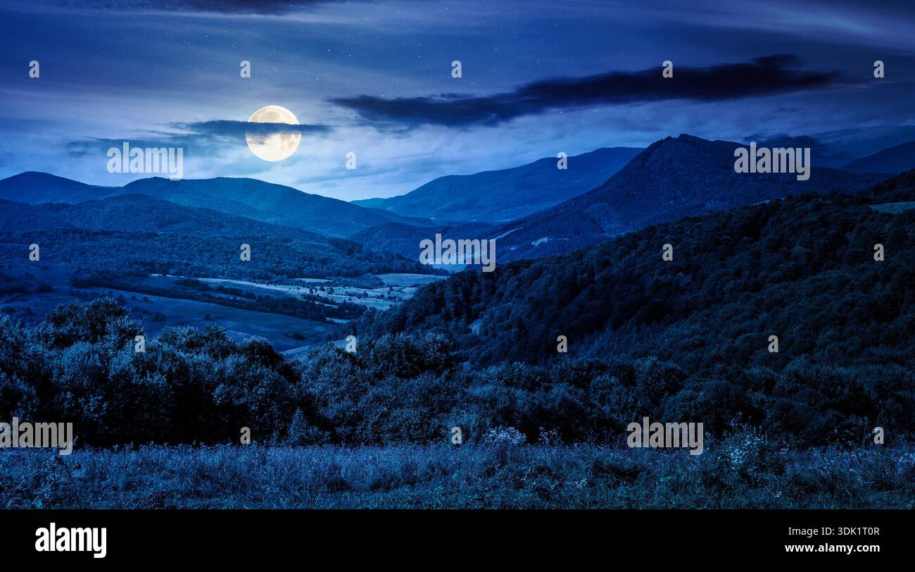 mountainous countryside scenery surrounded with forested rolling hills at night. rural area peaceful landscape with dark sky in full moon light. early Stock Photo