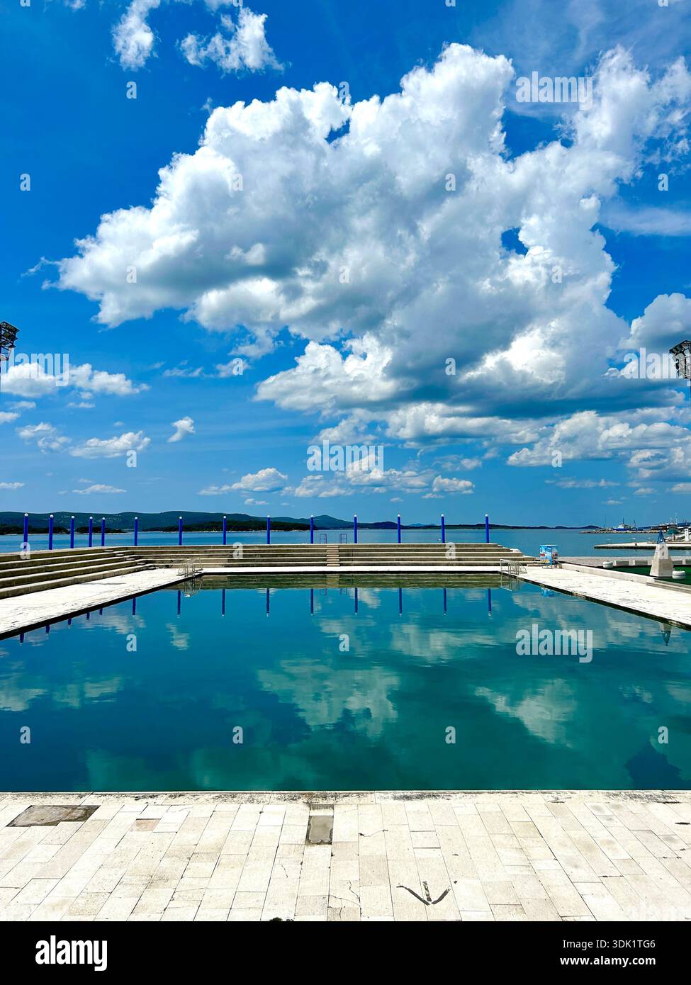 Outdoor public swimming pool overlooking the Adriatic Sea with blue summer sky an white fluffy clouds reflected in the water. - Smartphone Captured Stock Image
