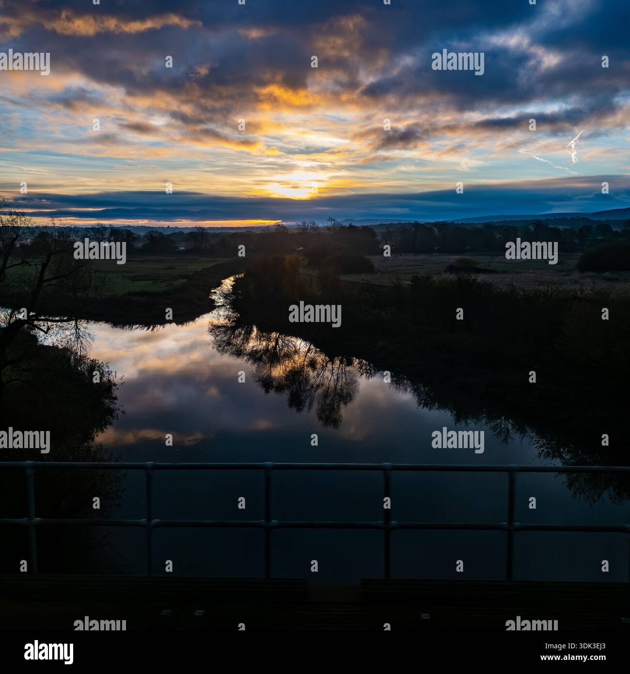 Stunning sunrise reflection of dramatic clouds, trees, and distant hills in a calm river viewed from a bridge railing in the countryside - Smartphone Captured Stock Image