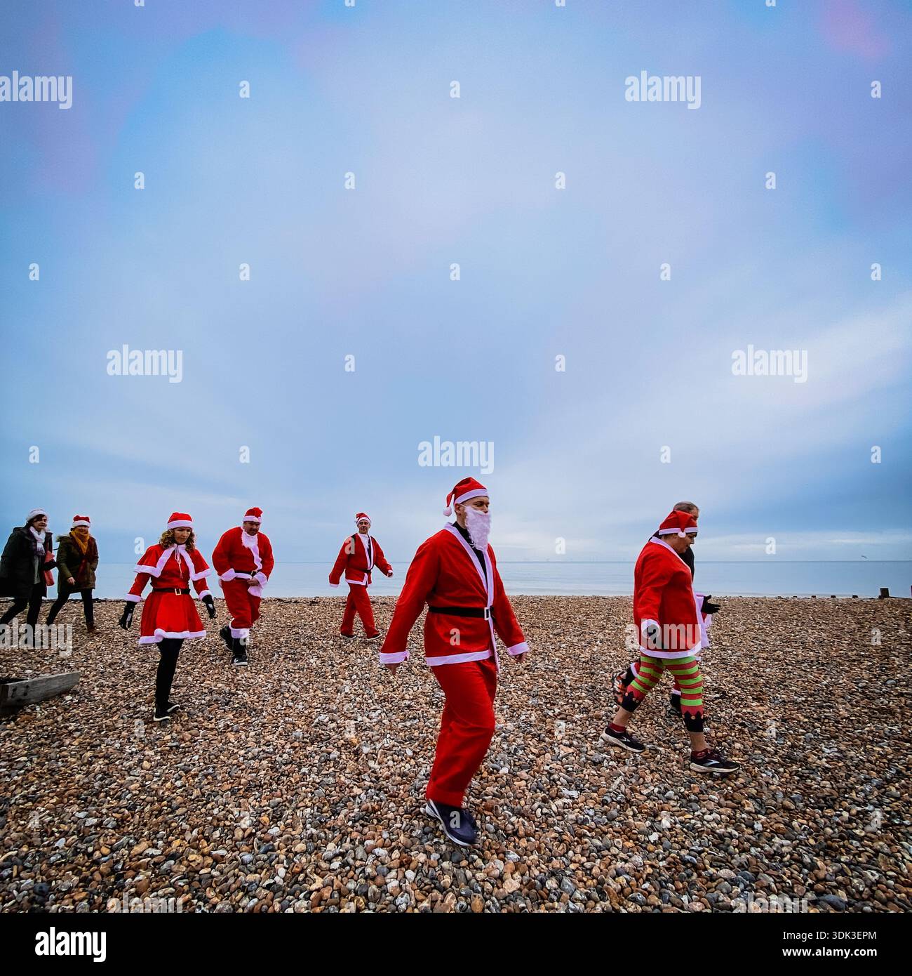 Diverse group in Santa Claus costumes walking and chatting on a pebbly beach under a vast cloudy sky, embodying joyful Christmas festivities by the se - Smartphone Captured Stock Image