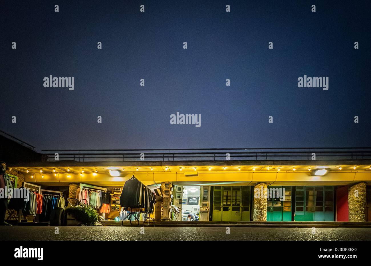 Vibrant East Beach Studios on Worthing Beach illuminated at night, showcasing colorful beach huts with hanging clothes and glowing lights against the - Smartphone Captured Stock Image