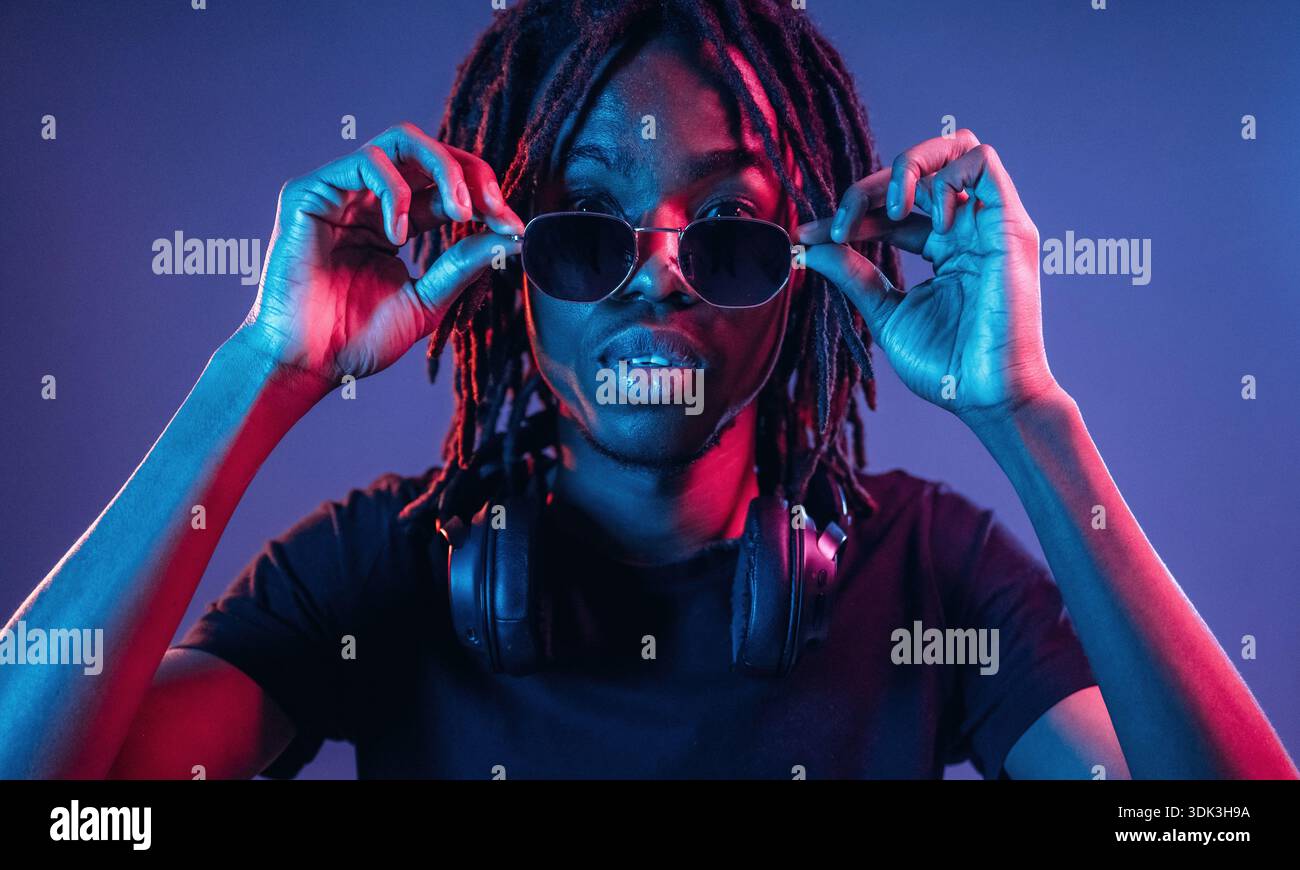 Touching sunglasses, front view. African American guy is studio with neon lighting. Stock Photo