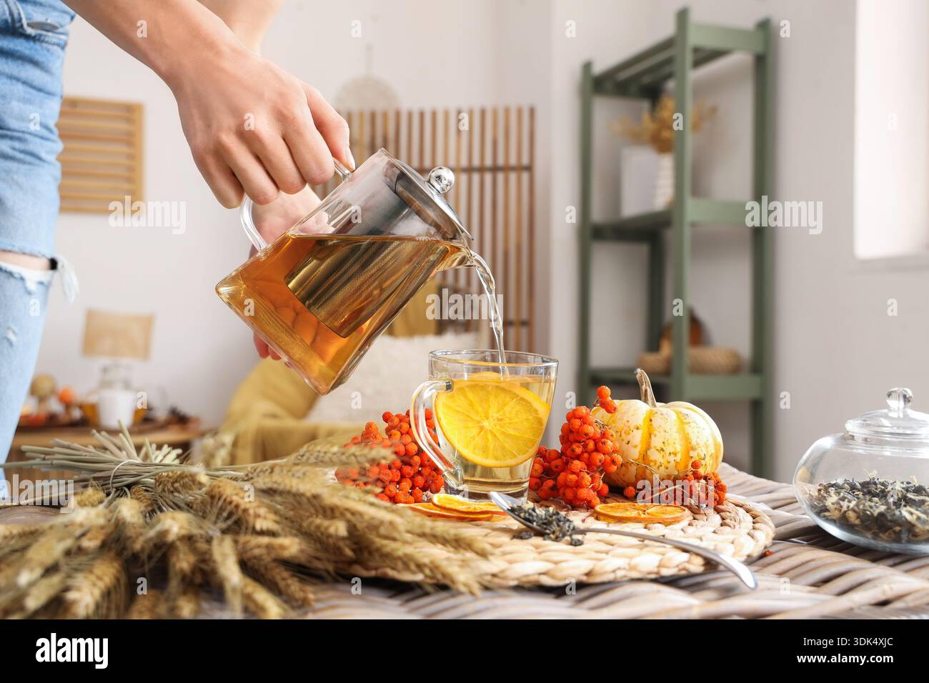 Female hand pouring tea from teapot into cup near bunch of wheat and rowan on table in room. Closeup Stock Photo