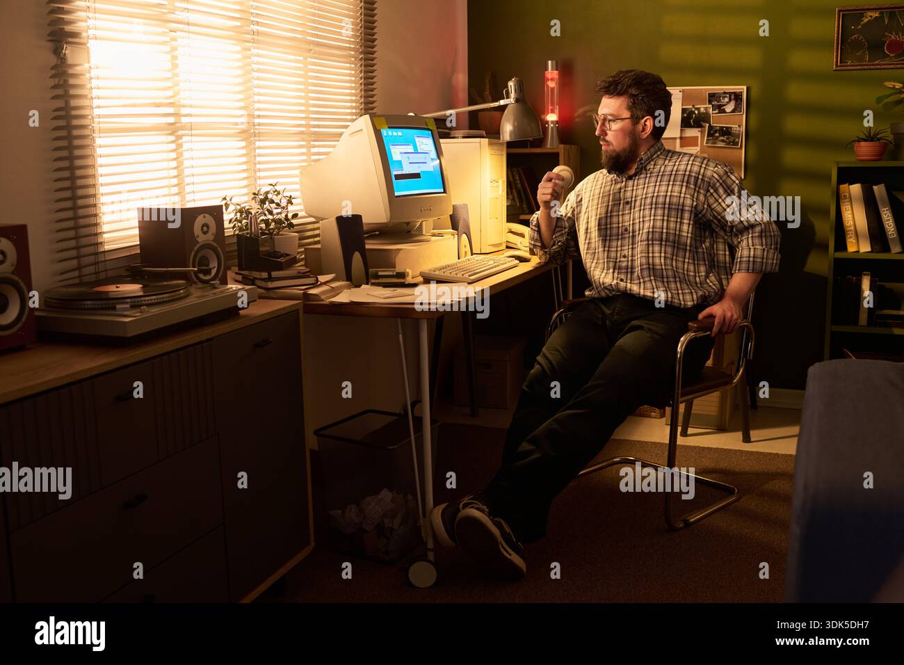 Caucasian man sitting at desk using vintage desktop computer while holding ball, looking at monitor in home office with retro electronics and decor visible in background Stock Photo