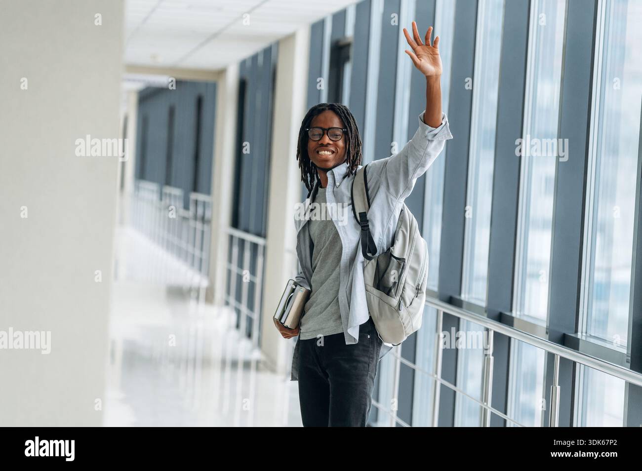 Bye hand gesture, smiling. Student with backpack is in the corridor. Stock Photo