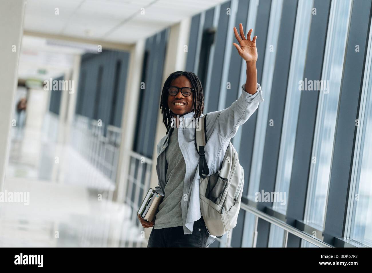 Bye hand gesture, smiling. Student with backpack is in the corridor. Stock Photo