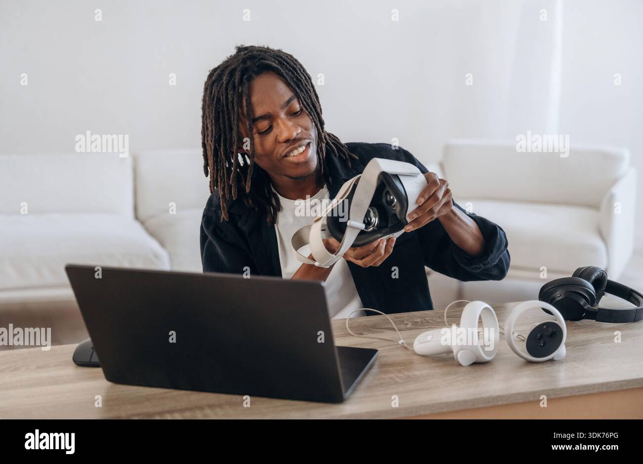 Wearing headset. Young man is sitting by table with laptop and virtual reality glasses. Stock Photo