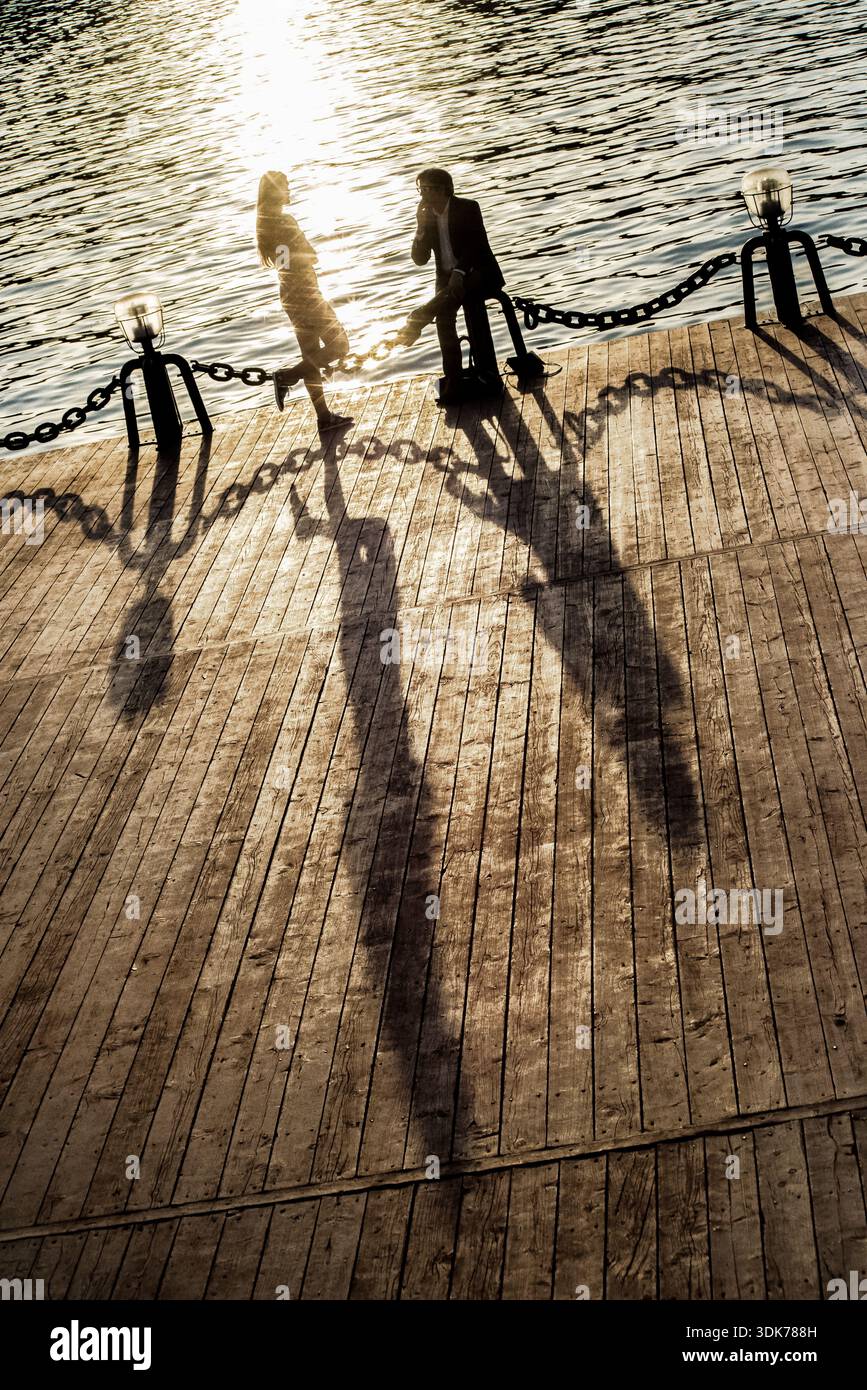 Two people standing by a chain barrier near water, casting long shadows on a paved surface. Stock Photo