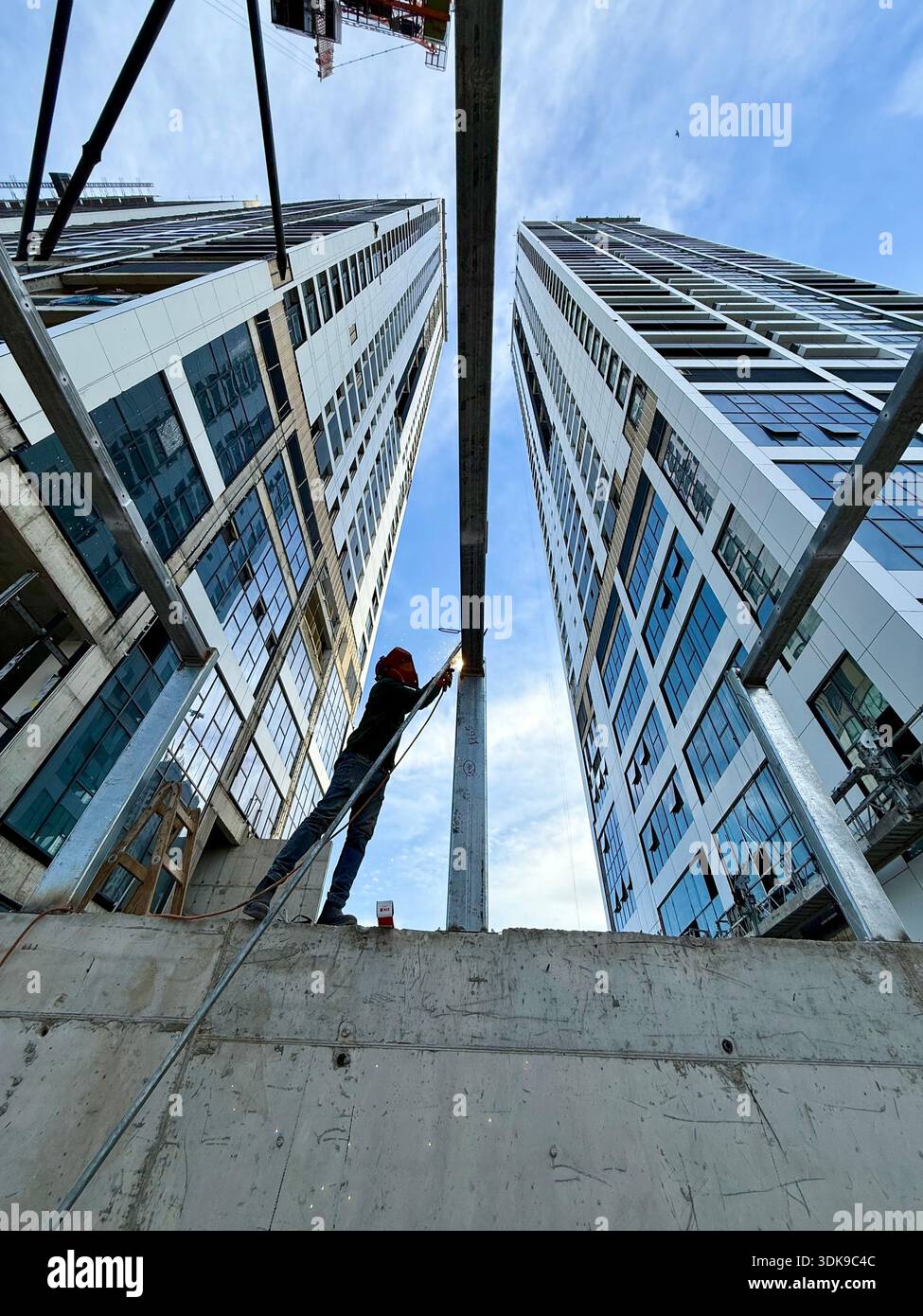 Construction worker on a ladder welding steel beams at a high-rise building site. Low-angle view of industrial labor against a modern skyscraper. - Smartphone Captured Stock Image