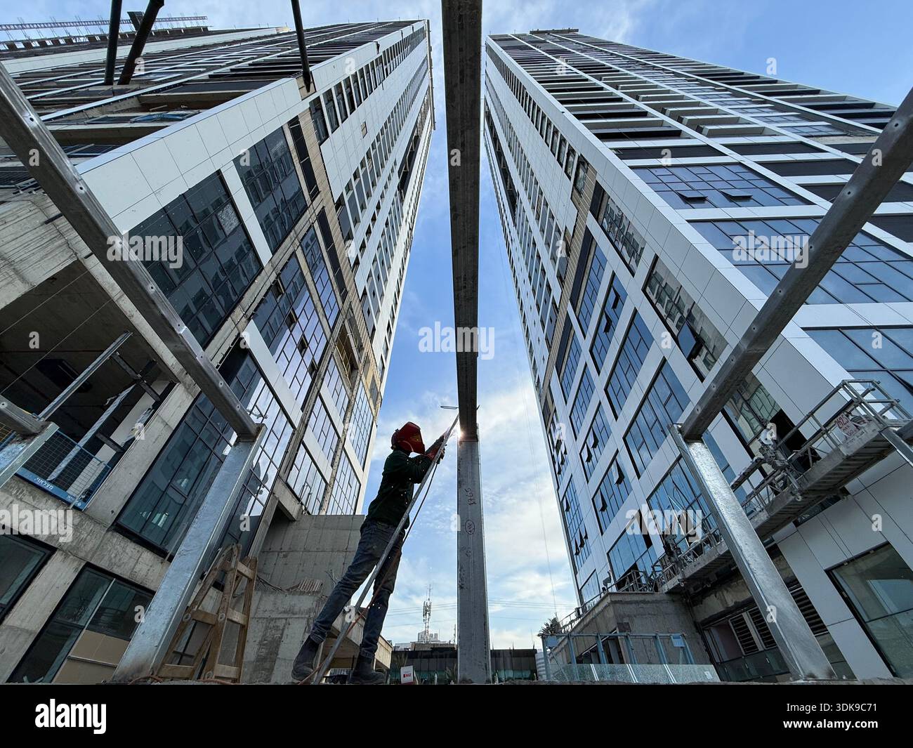 Professional welder in safety mask welding steel beams on a high-rise construction site. Low-angle view with industrial crane and modern buildings. - Smartphone Captured Stock Image