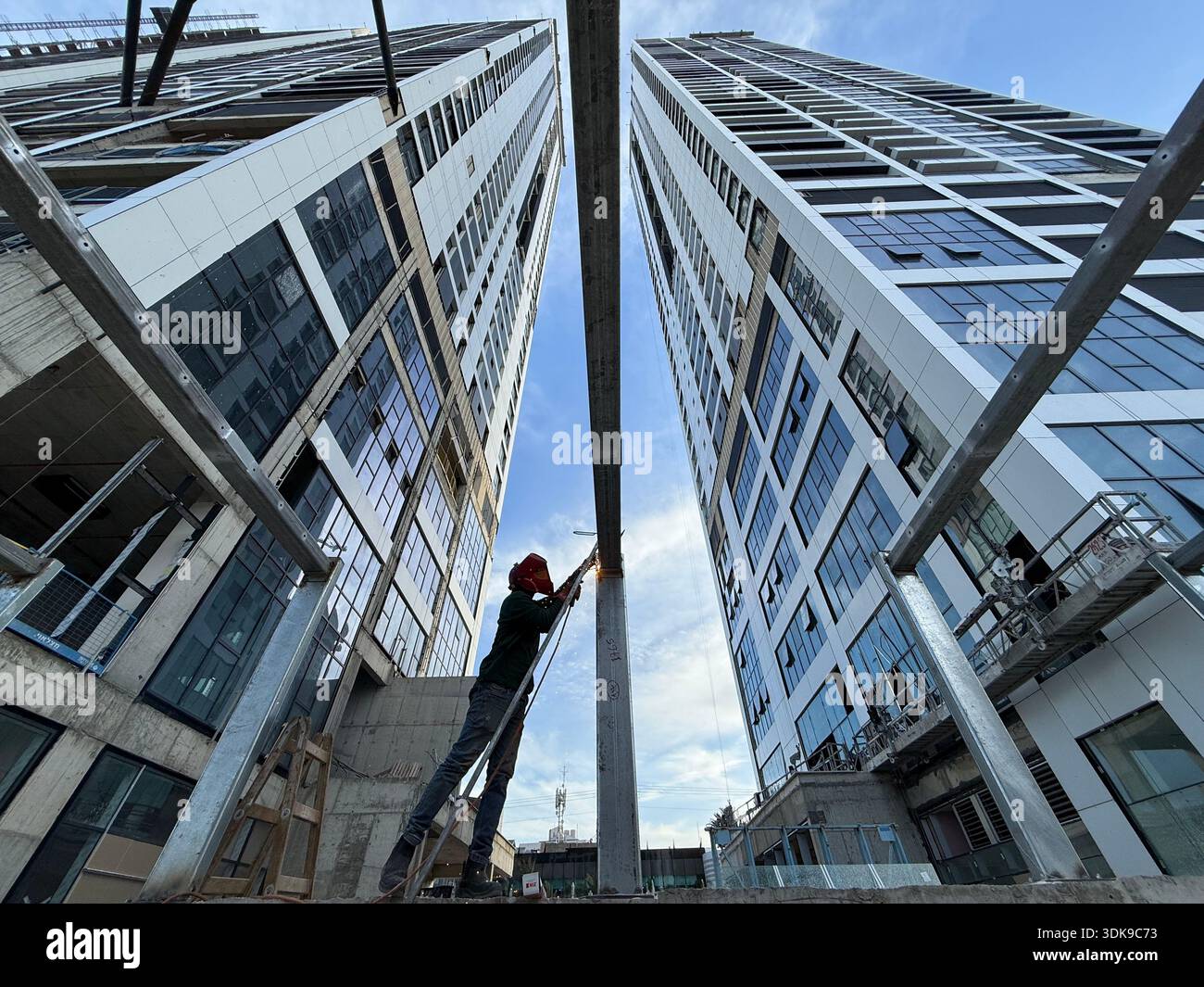 Professional welder in safety mask welding steel beams on a high-rise construction site. Low-angle view with industrial crane and modern buildings. - Smartphone Captured Stock Image