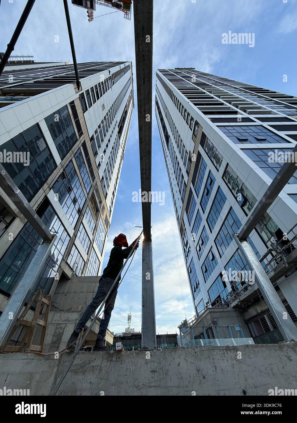 Professional welder in safety mask welding steel beams on a high-rise construction site. Low-angle view with industrial crane and modern buildings. - Smartphone Captured Stock Image
