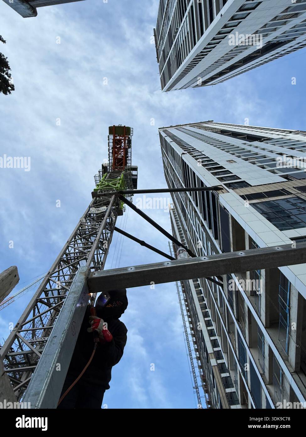 Professional welder in safety mask welding steel beams on a high-rise construction site. Low-angle view with industrial crane and modern buildings. - Smartphone Captured Stock Image