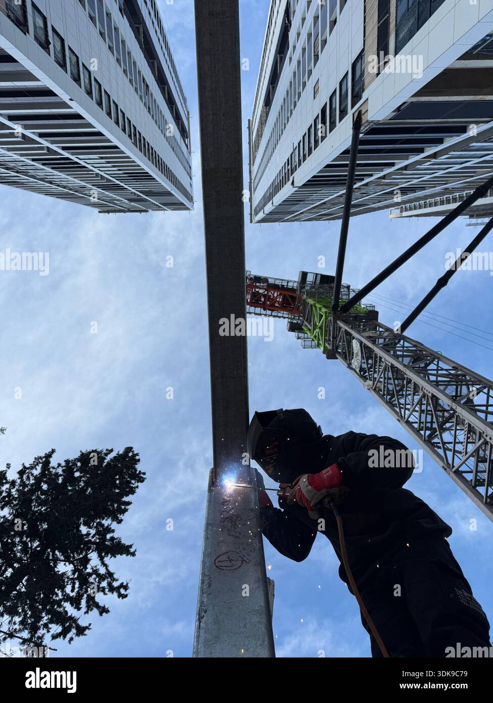 Professional welder in safety mask welding steel beams on a high-rise construction site. Low-angle view with industrial crane and modern buildings. - Smartphone Captured Stock Image