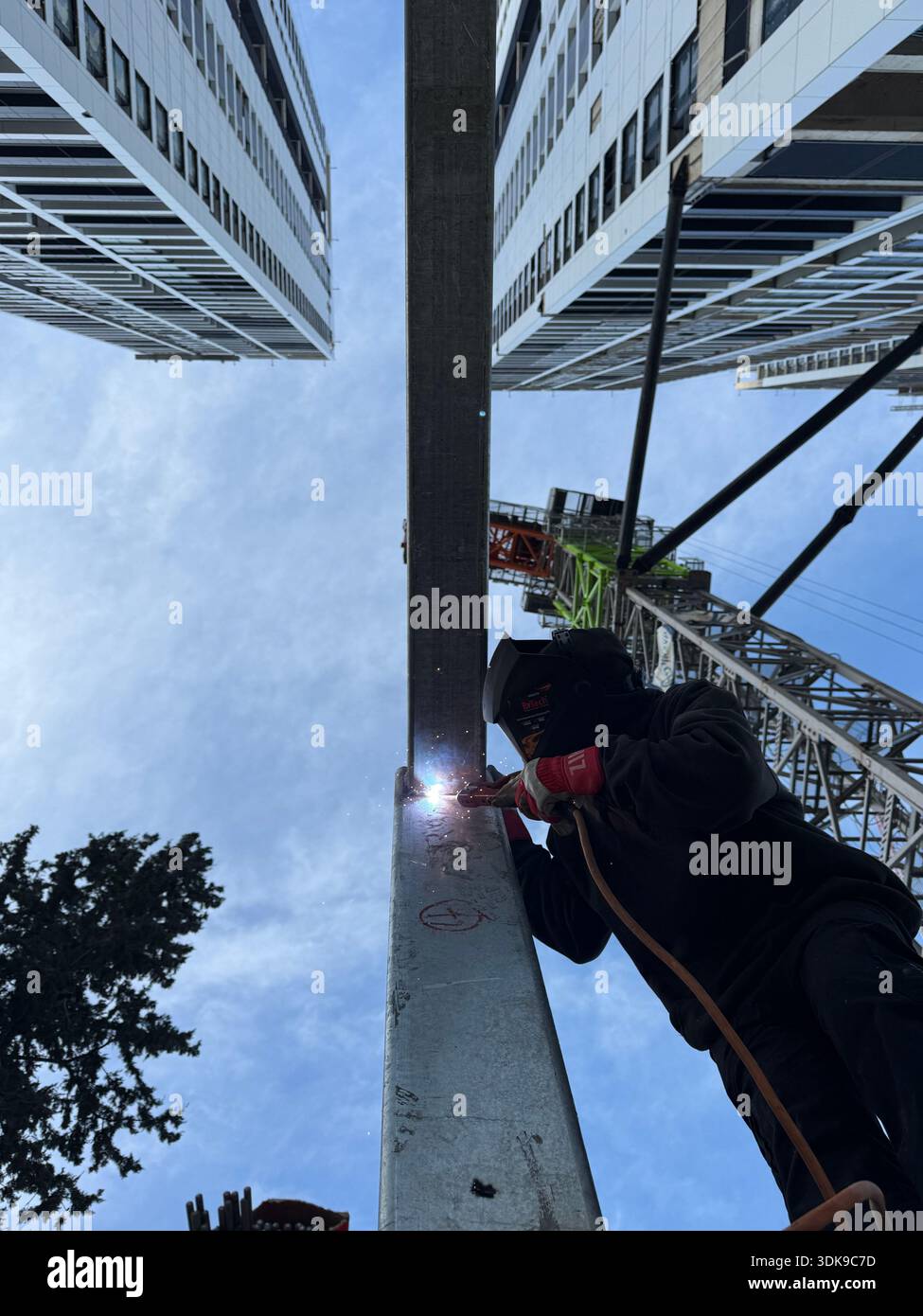 Professional welder in safety mask welding steel beams on a high-rise construction site. Low-angle view with industrial crane and modern buildings. - Smartphone Captured Stock Image