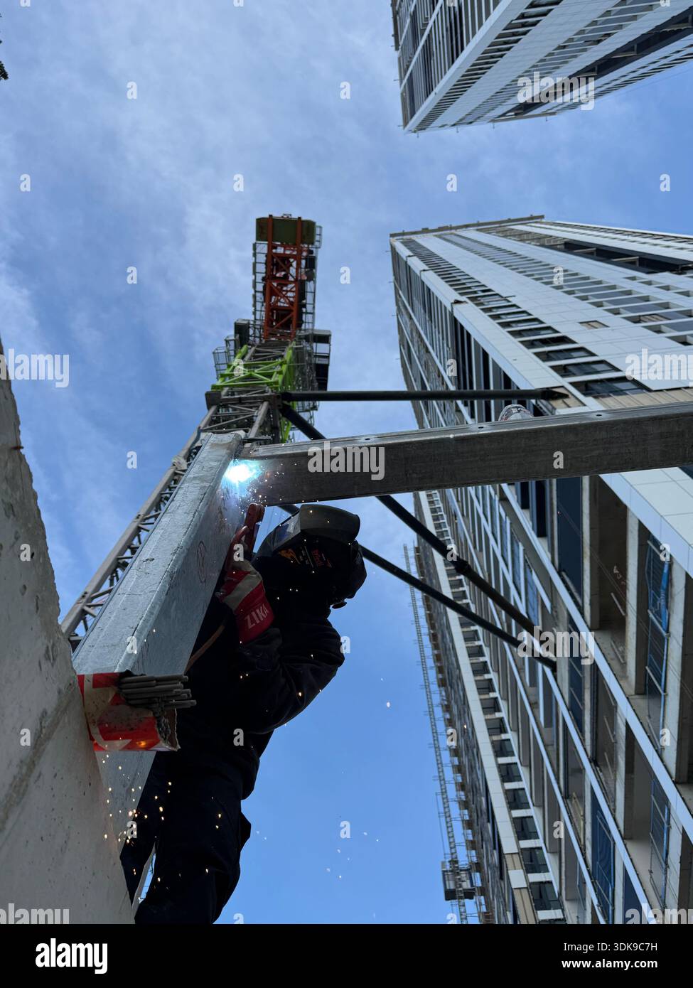 Professional welder in safety mask welding steel beams on a high-rise construction site. Low-angle view with industrial crane and modern buildings. - Smartphone Captured Stock Image