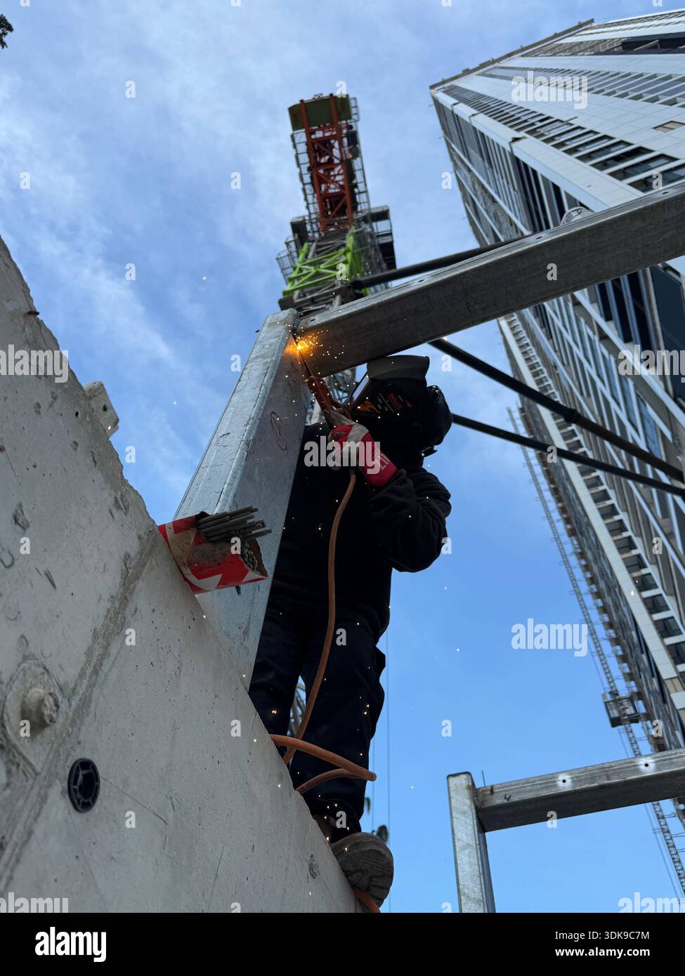Professional welder in safety mask welding steel beams on a high-rise construction site. Low-angle view with industrial crane and modern buildings. - Smartphone Captured Stock Image
