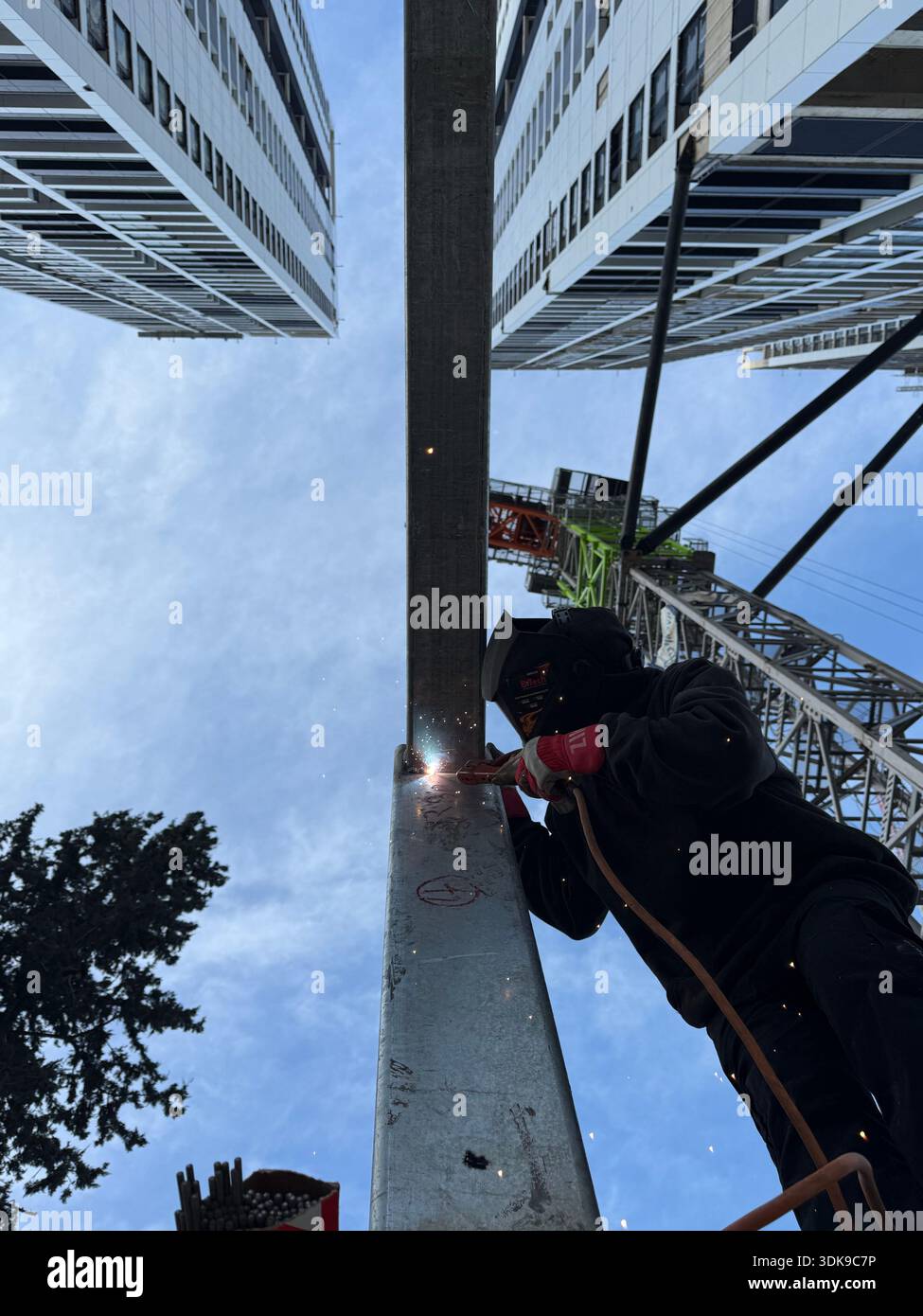 Professional welder in safety mask welding steel beams on a high-rise construction site. Low-angle view with industrial crane and modern buildings. - Smartphone Captured Stock Image