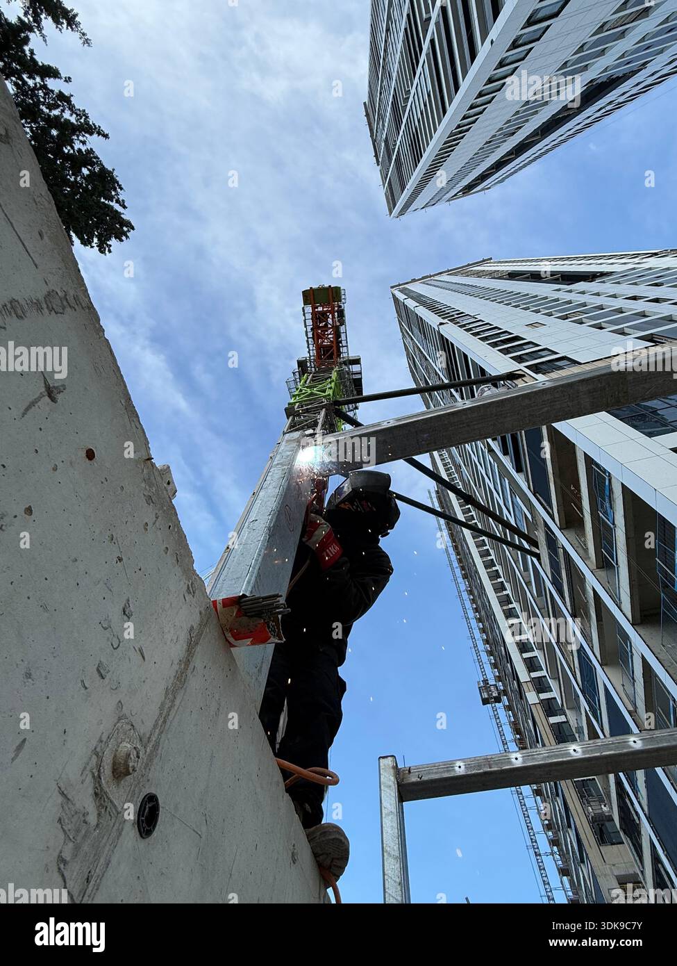 Professional welder in safety mask welding steel beams on a high-rise construction site. Low-angle view with industrial crane and modern buildings. - Smartphone Captured Stock Image