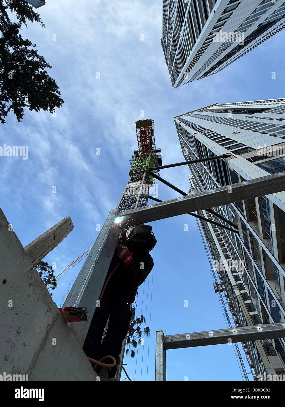 Professional welder in safety mask welding steel beams on a high-rise construction site. Low-angle view with industrial crane and modern buildings. - Smartphone Captured Stock Image