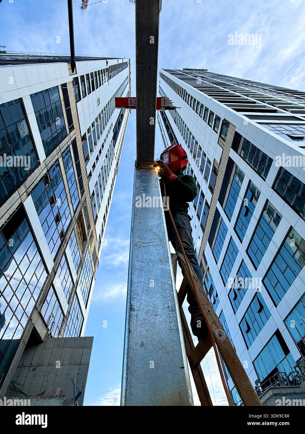 Professional welder in safety mask welding steel beams on a high-rise construction site. Low-angle view with industrial crane and modern buildings. - Smartphone Captured Stock Image