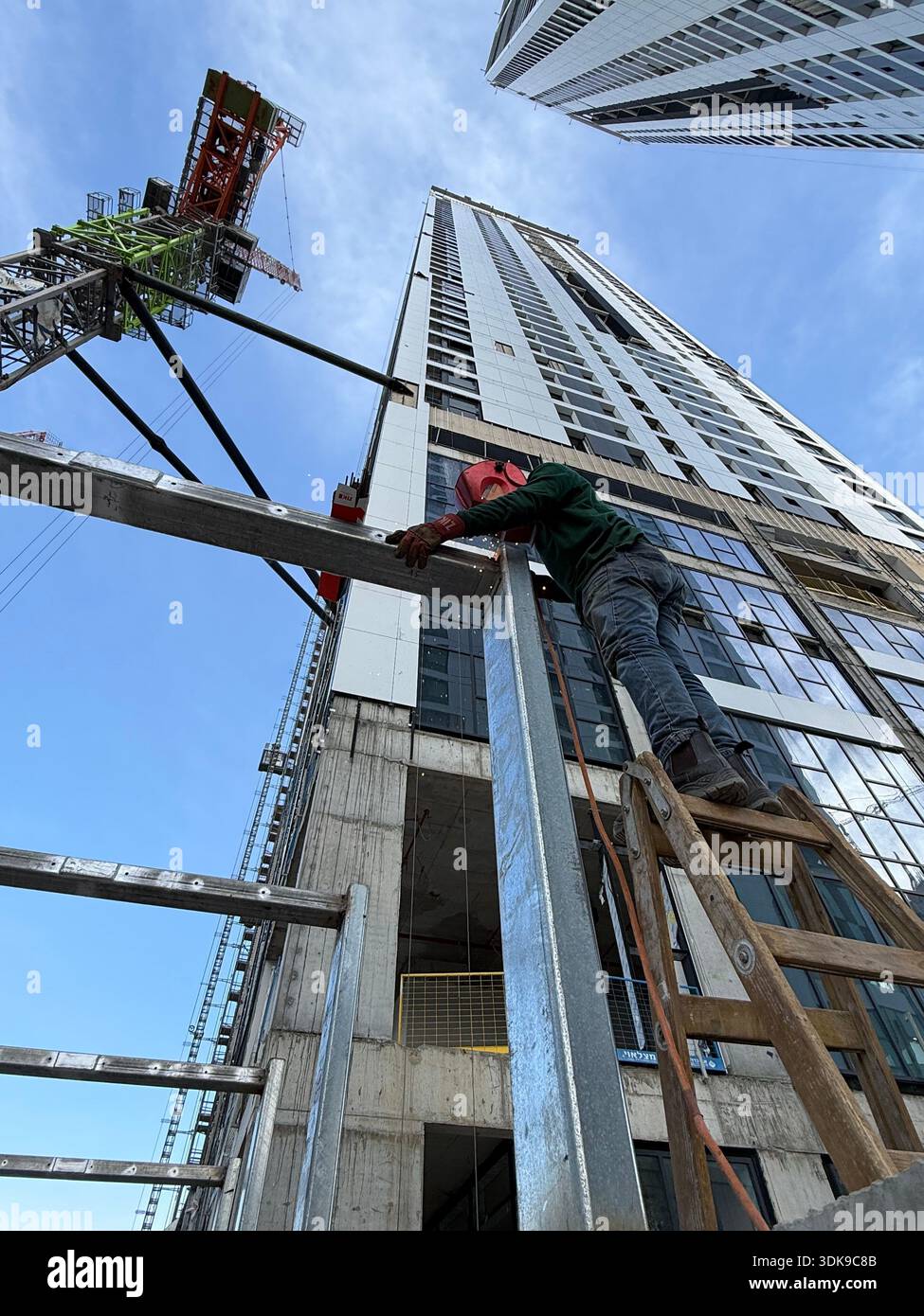 Professional welder in safety mask welding steel beams on a high-rise construction site. Low-angle view with industrial crane and modern buildings. - Smartphone Captured Stock Image