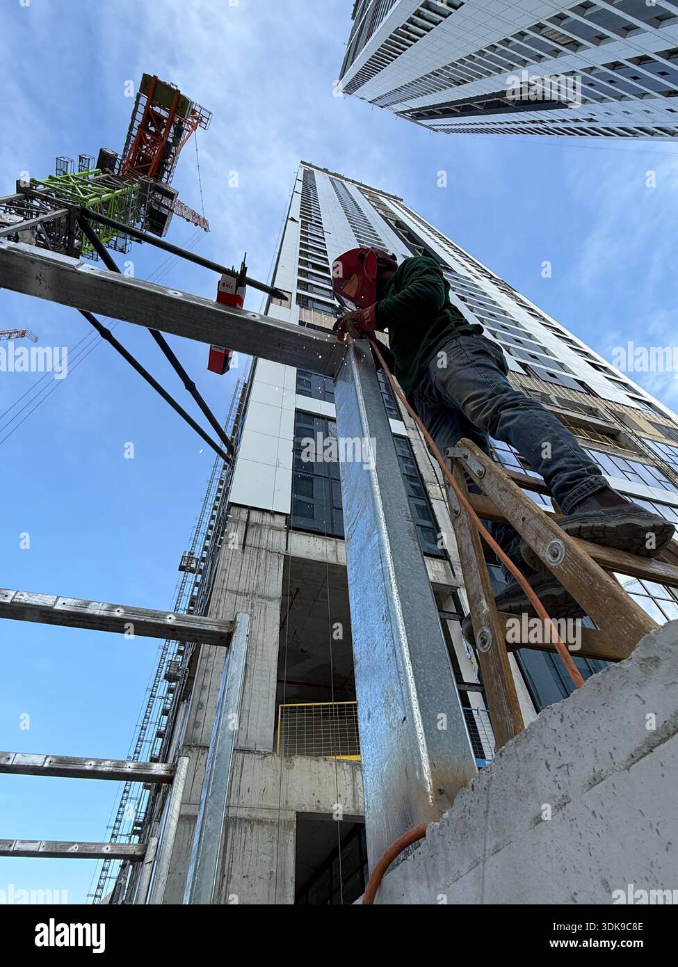 Professional welder in safety mask welding steel beams on a high-rise construction site. Low-angle view with industrial crane and modern buildings. - Smartphone Captured Stock Image