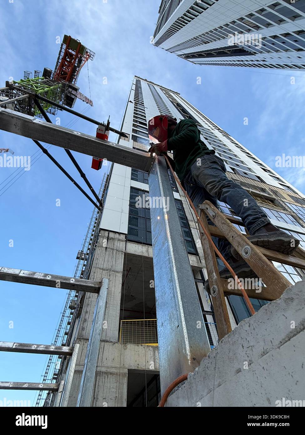 Professional welder in safety mask welding steel beams on a high-rise construction site. Low-angle view with industrial crane and modern buildings. - Smartphone Captured Stock Image