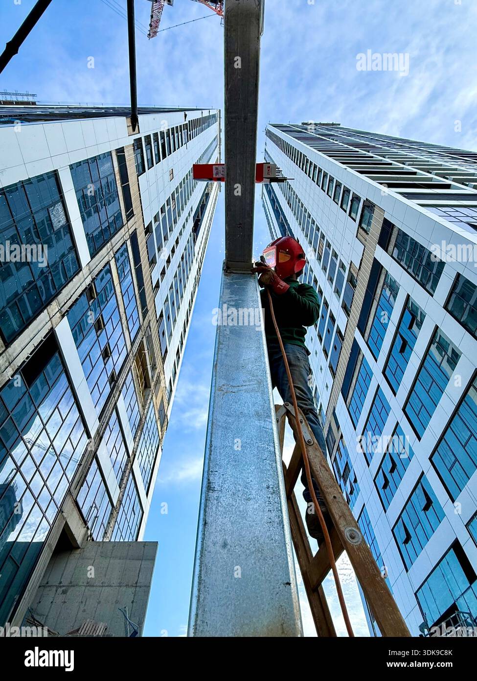 Professional welder in safety mask welding steel beams on a high-rise construction site. Low-angle view with industrial crane and modern buildings. - Smartphone Captured Stock Image