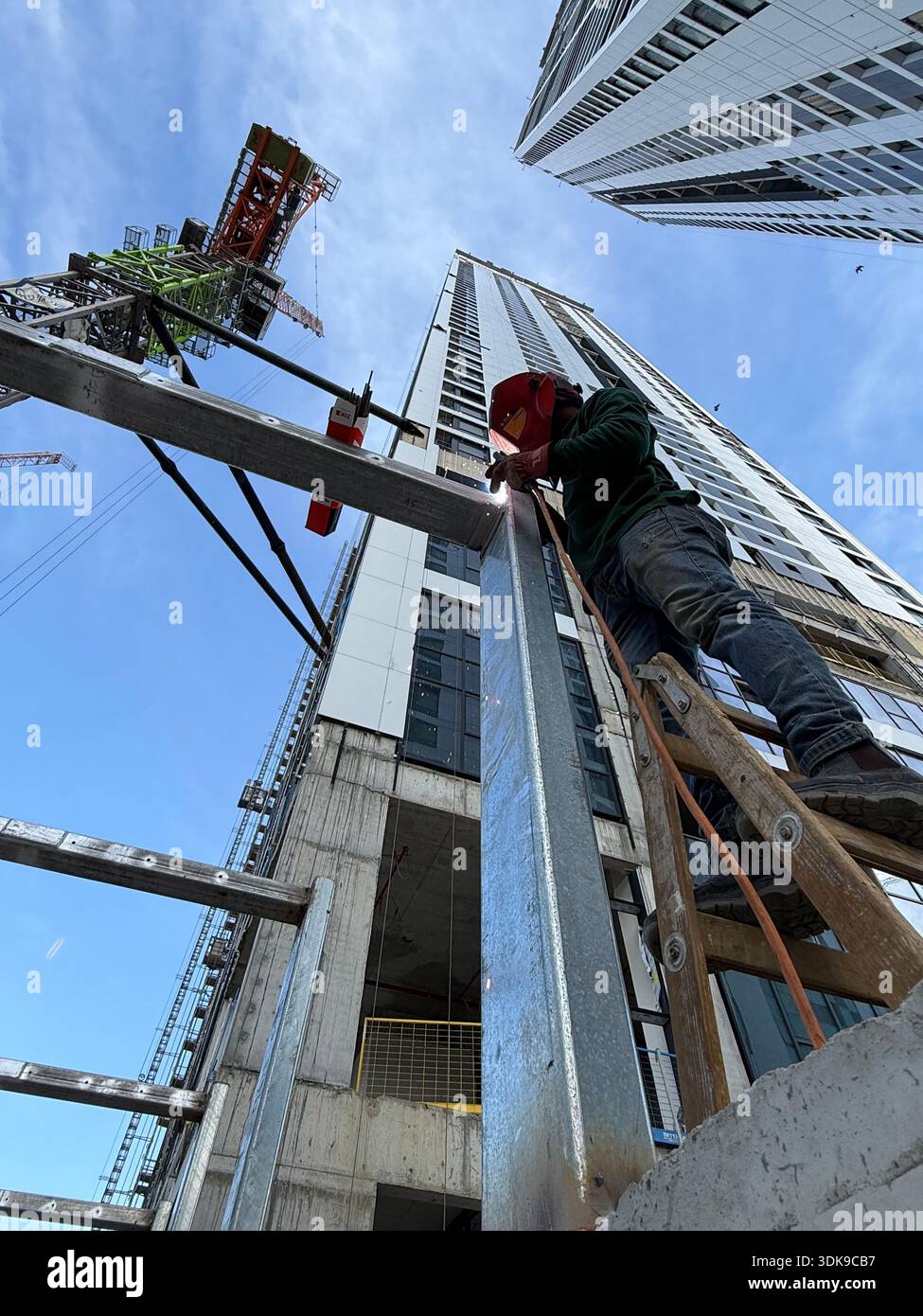 Professional welder in safety mask welding steel beams on a high-rise construction site. Low-angle view with industrial crane and modern buildings. - Smartphone Captured Stock Image