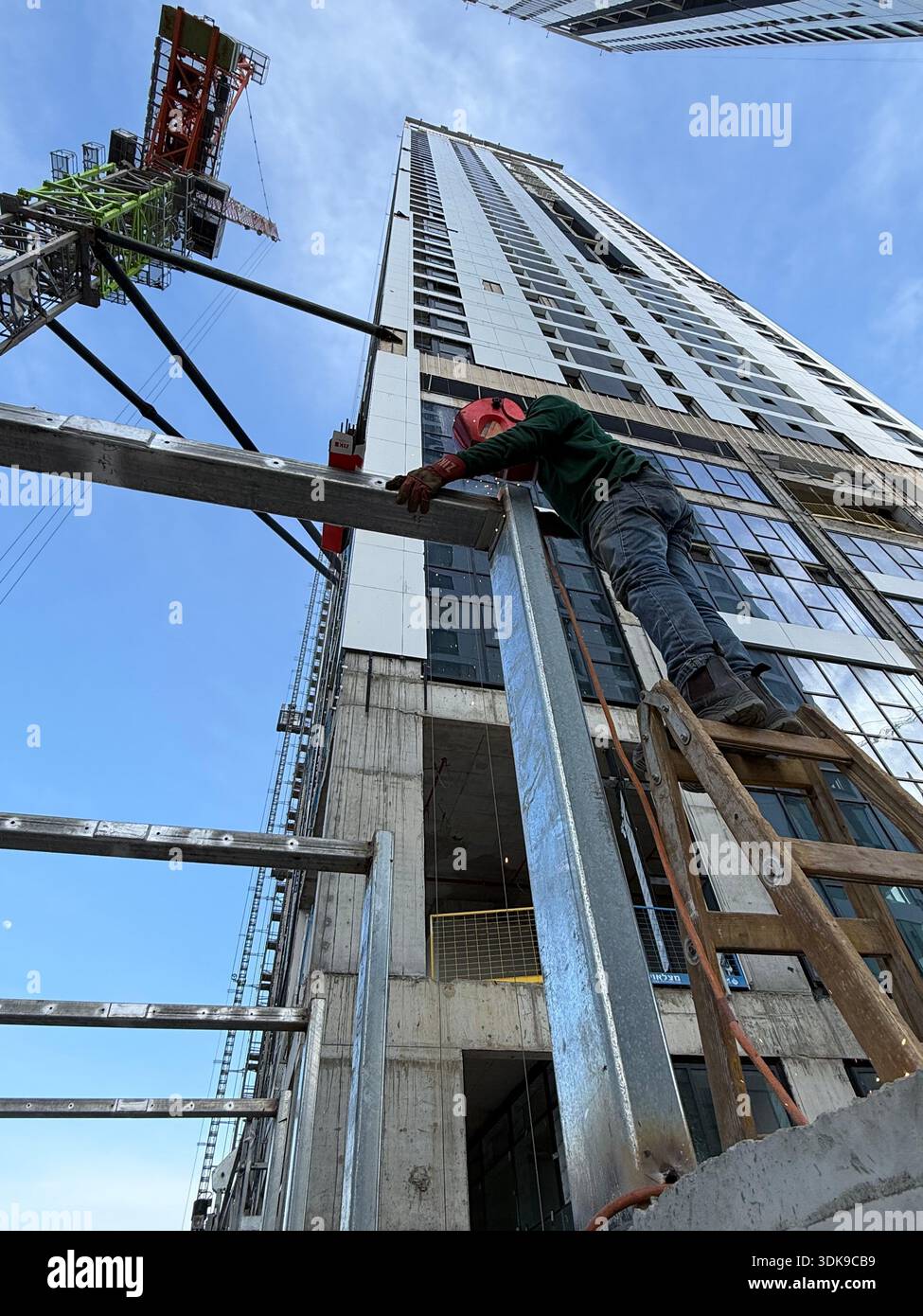 Professional welder in safety mask welding steel beams on a high-rise construction site. Low-angle view with industrial crane and modern buildings. - Smartphone Captured Stock Image