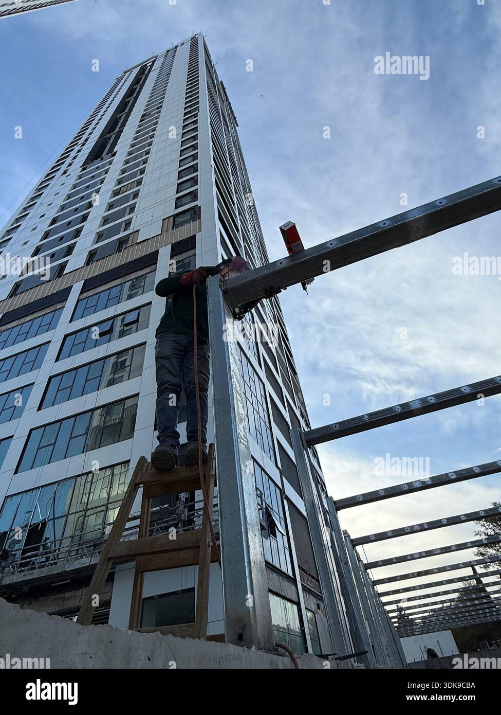 Construction worker on a ladder welding steel beams at a high-rise building site. Low-angle view of industrial labor against a modern skyscraper. - Smartphone Captured Stock Image