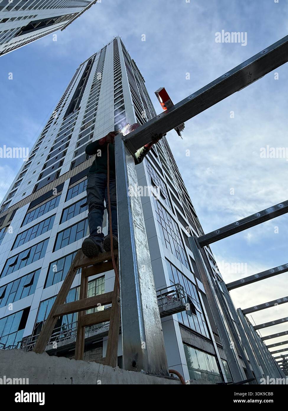 Construction worker on a ladder welding steel beams at a high-rise building site. Low-angle view of industrial labor against a modern skyscraper. - Smartphone Captured Stock Image