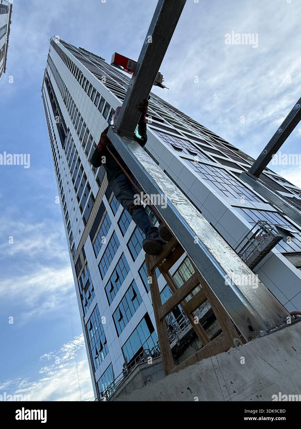 Construction worker on a ladder welding steel beams at a high-rise building site. Low-angle view of industrial labor against a modern skyscraper. - Smartphone Captured Stock Image