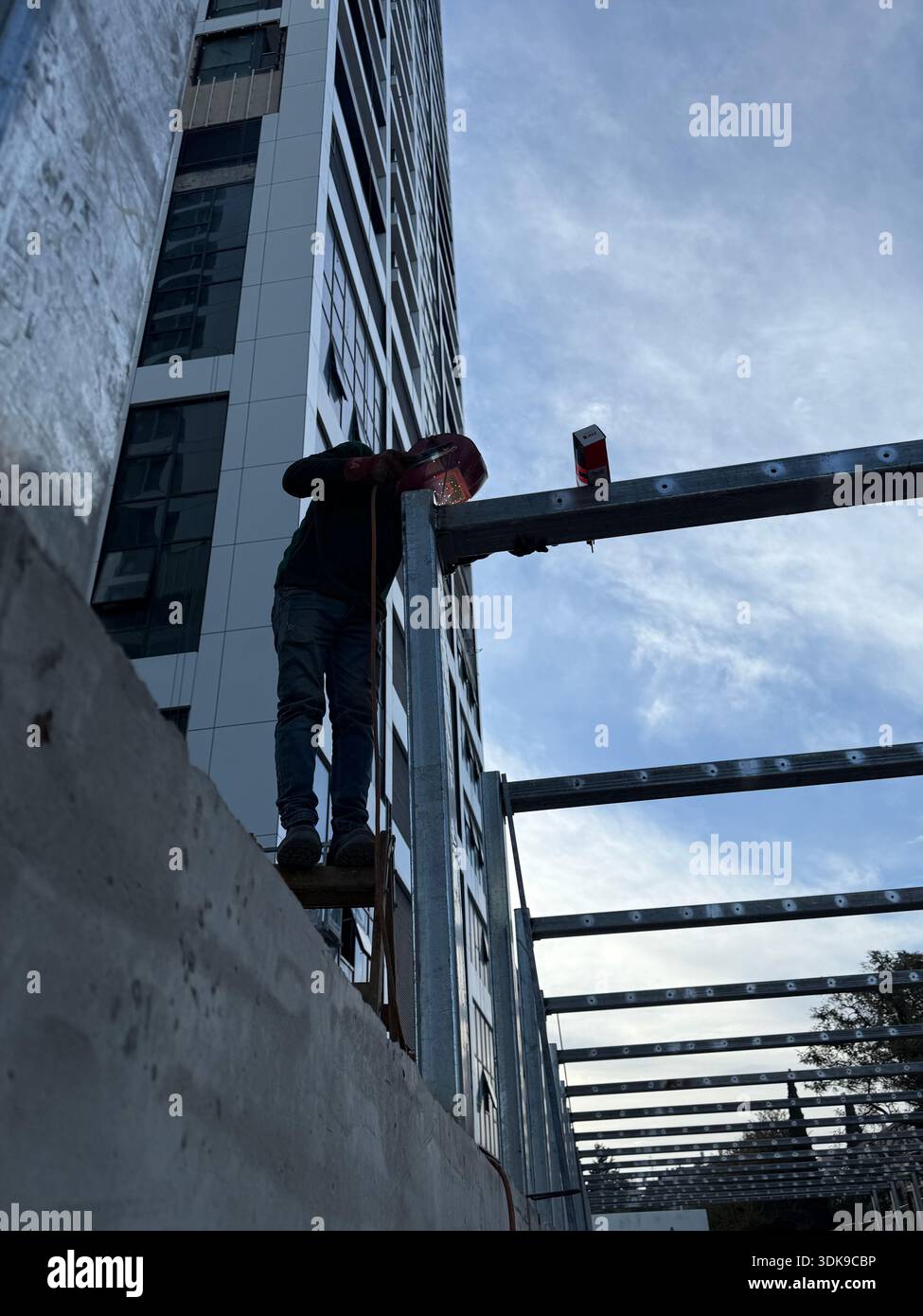 Construction worker on a ladder welding steel beams at a high-rise building site. Low-angle view of industrial labor against a modern skyscraper. - Smartphone Captured Stock Image